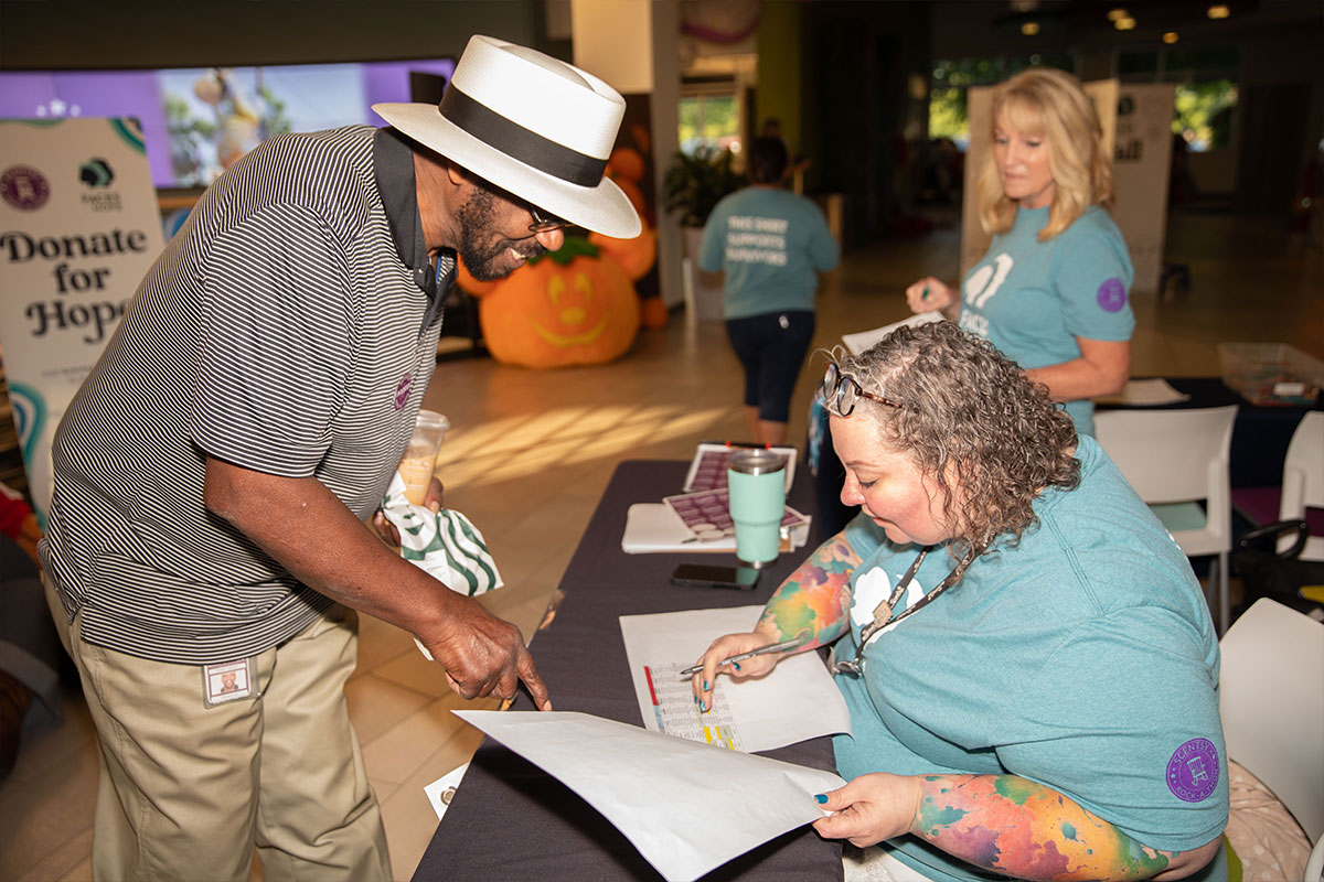 woman in teal tee shirt helping a man with a white sun hat sign up for rockathon